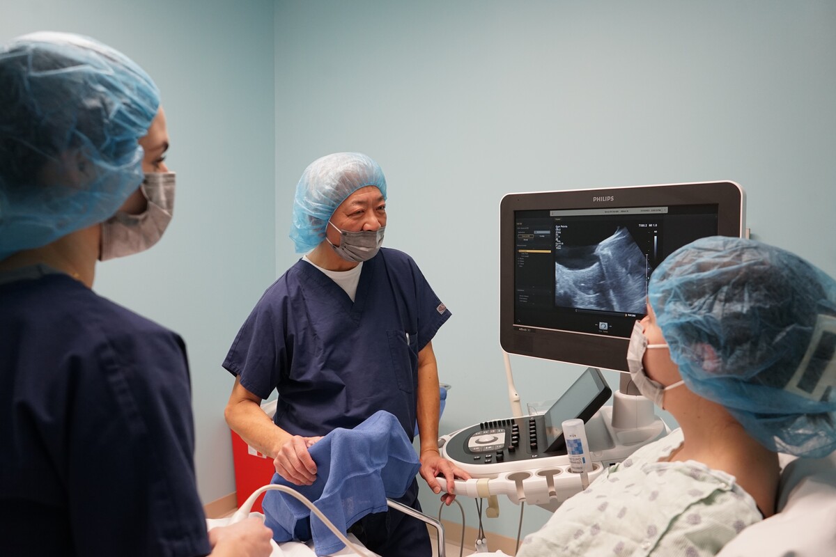 A doctor in scrubs speaks to a patient in an exam gown with a screen displaying an ultrasound image in between them