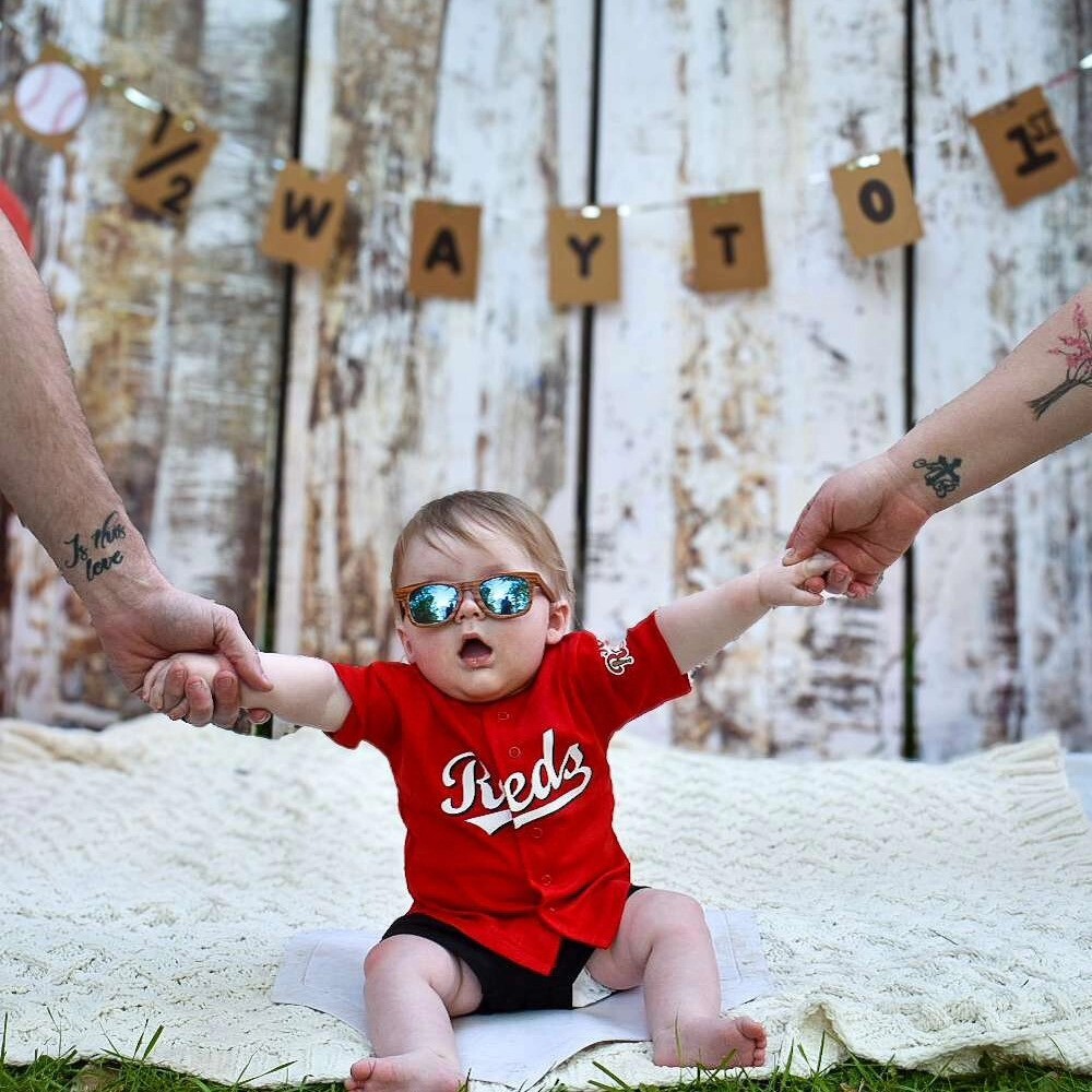 A baby wearing sunglasses and a Cincinnati reds shirt