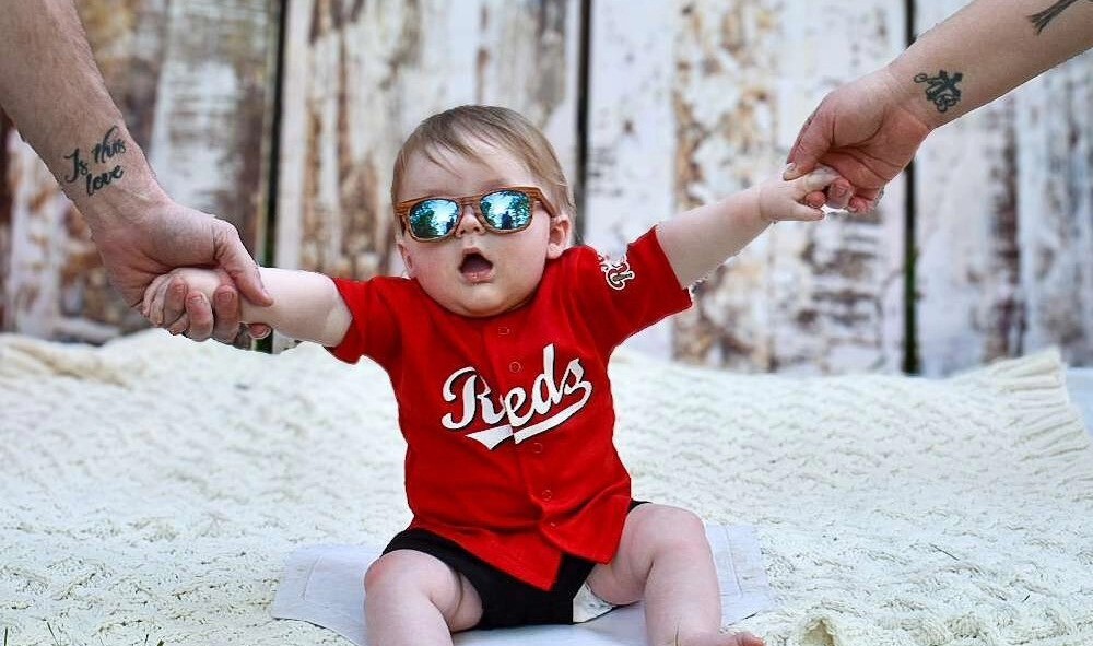 A baby wearing sunglasses and a Cincinnati reds shirt
